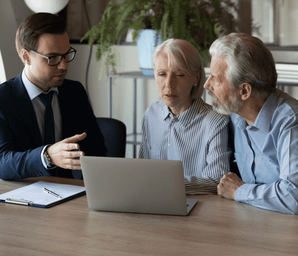 A man in a suit instructs an elderly couple using a laptop, seated at a table with a clipboard in a modern office setting. - Home Instead