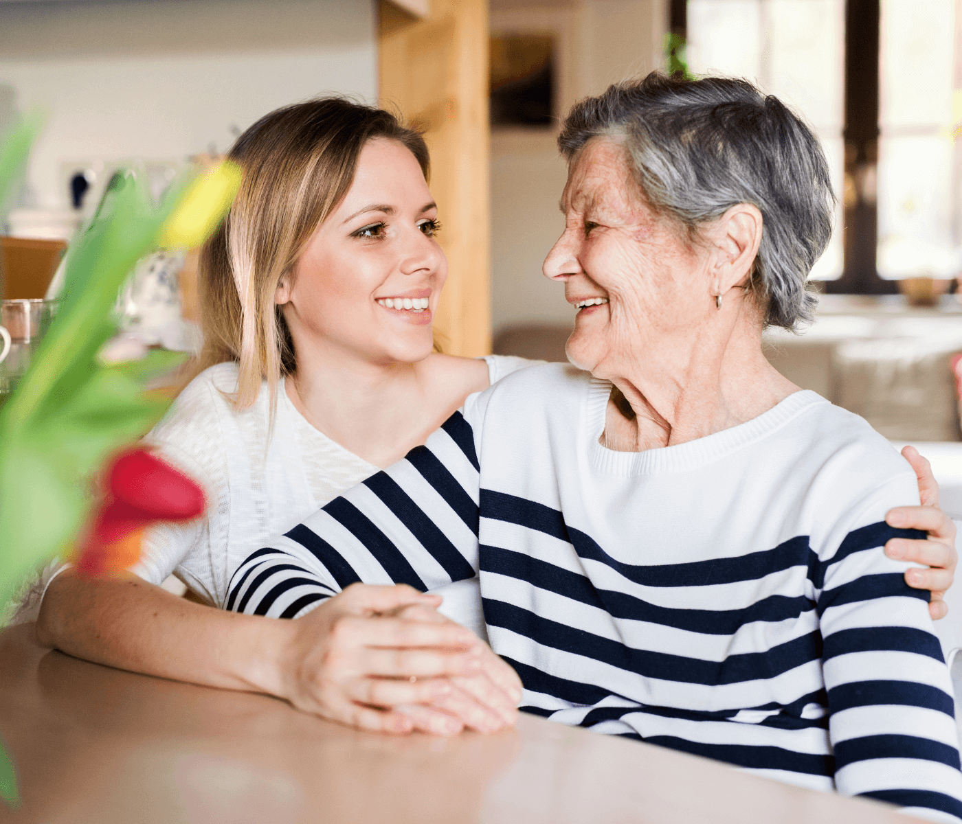 A young woman embraces an elderly woman, both smiling warmly, in a bright room with flowers in the foreground. - Home Instead