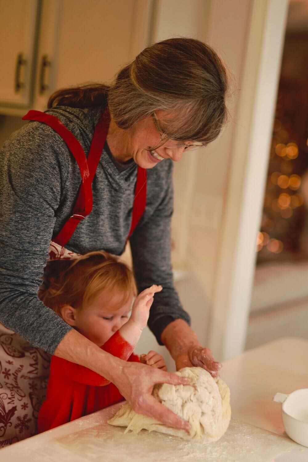 Elderly woman and young child kneading dough together in a warmly lit kitchen, both smiling and enjoying the activity. - Home Instead
