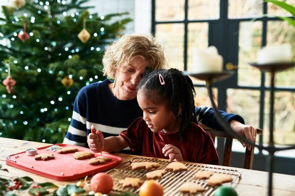 An adult and child decorate holiday cookies together at a table, near a Christmas tree with lights and ornaments. - Home Instead