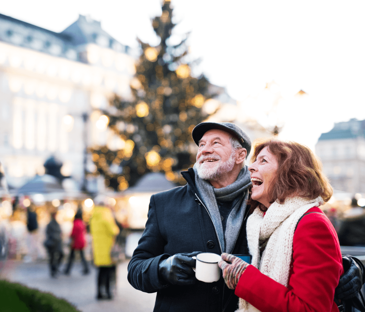 An elderly couple, smiling and holding a cup, stands in front of a festive city square with a decorated Christmas tree. - Home Instead