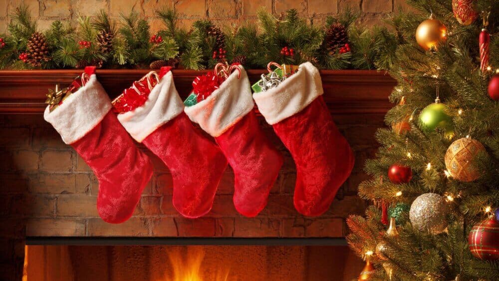 Four red Christmas stockings filled with gifts hanging over a decorated fireplace with a lit Christmas tree nearby. - Home Instead
