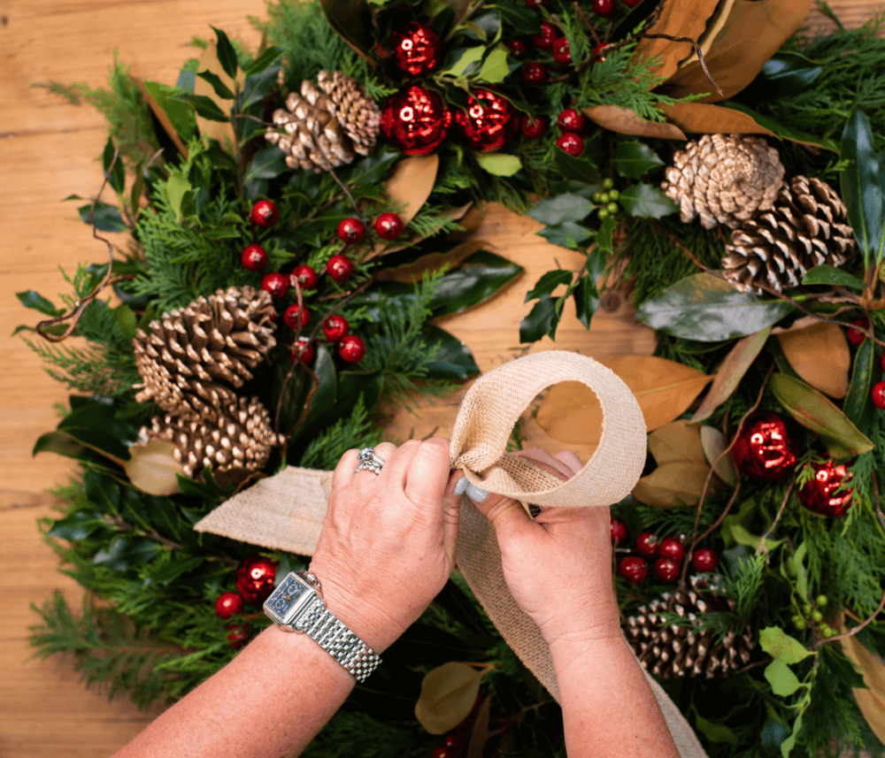 Hands tying a burlap ribbon on a festive holiday wreath decorated with pinecones, red berries, and greenery. - Home Instead