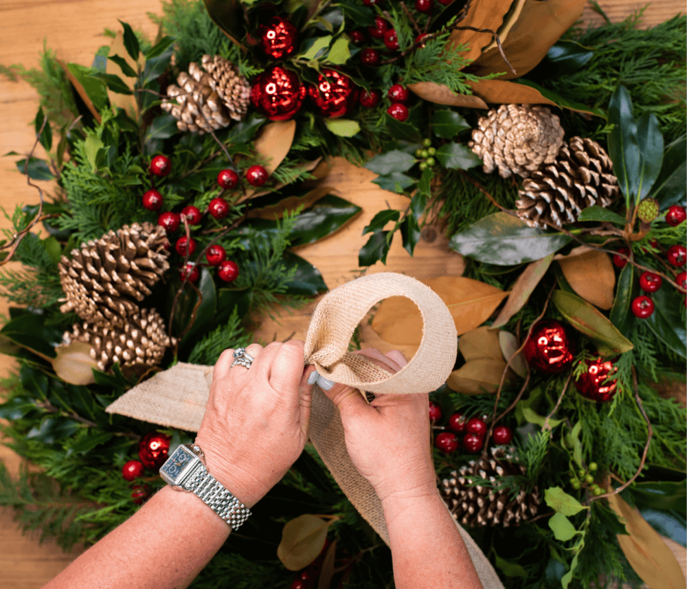 Hands tying a burlap ribbon on a festive Christmas wreath decorated with pinecones and red ornaments. - Home Instead