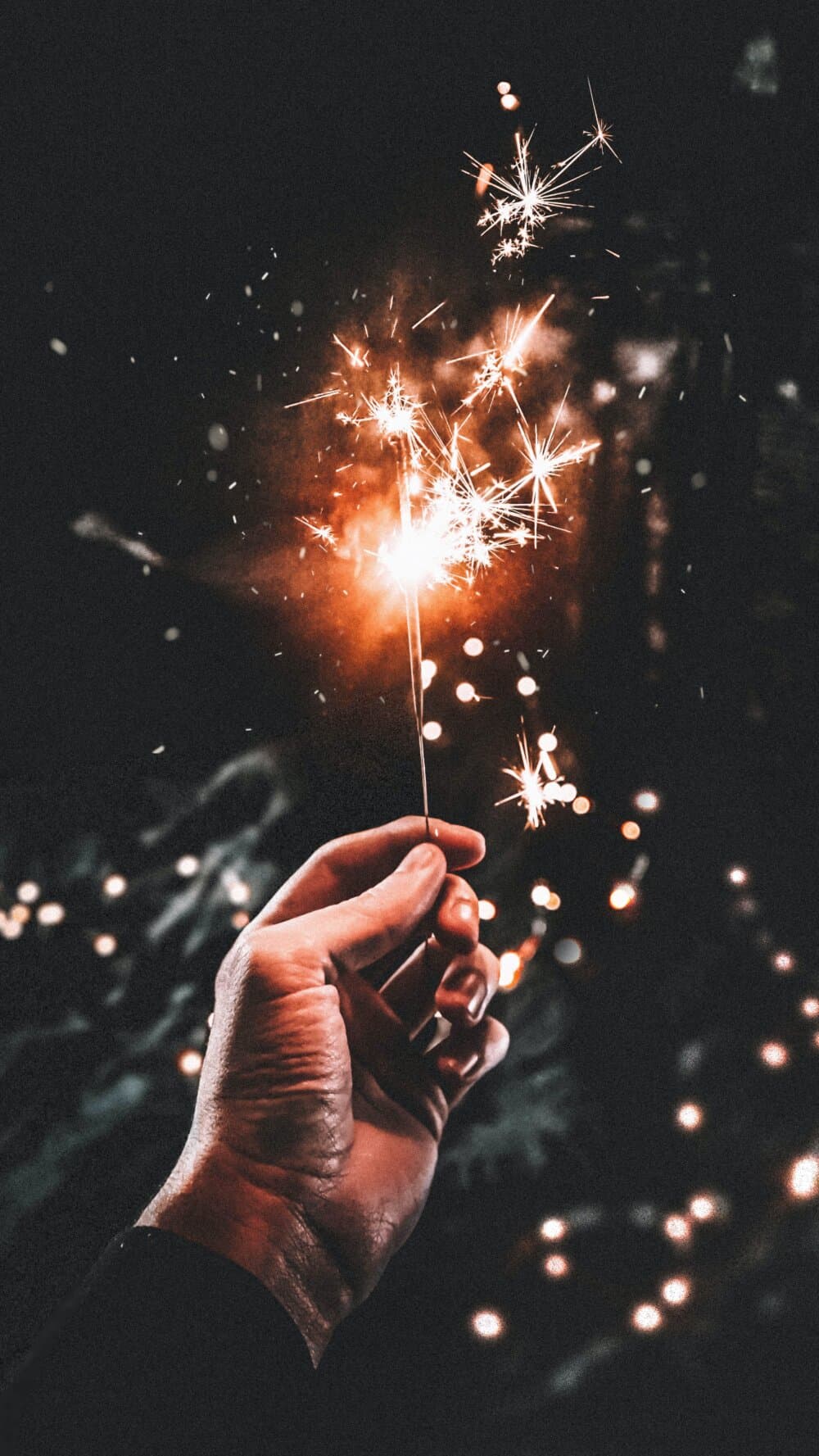 Hand holding a lit sparkler with glowing sparks against a dark background. - Home Instead