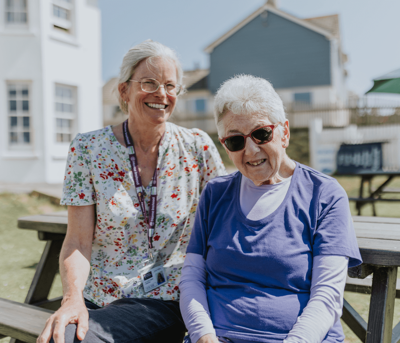 Two smiling elderly women sit on a picnic bench outside on a sunny day, one in a floral shirt and one in a blue shirt. - Home Instead