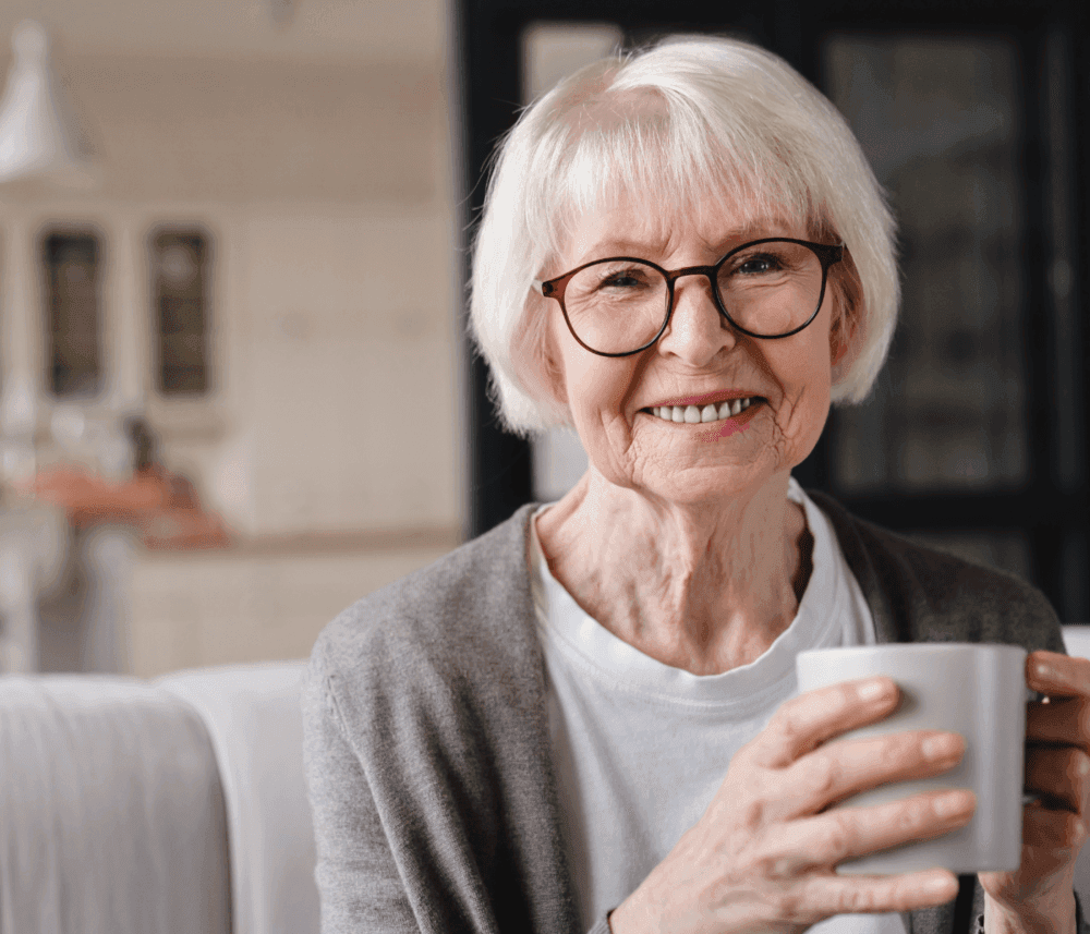 Elderly woman with white hair and glasses smiles while holding a white mug indoors. - Home Instead