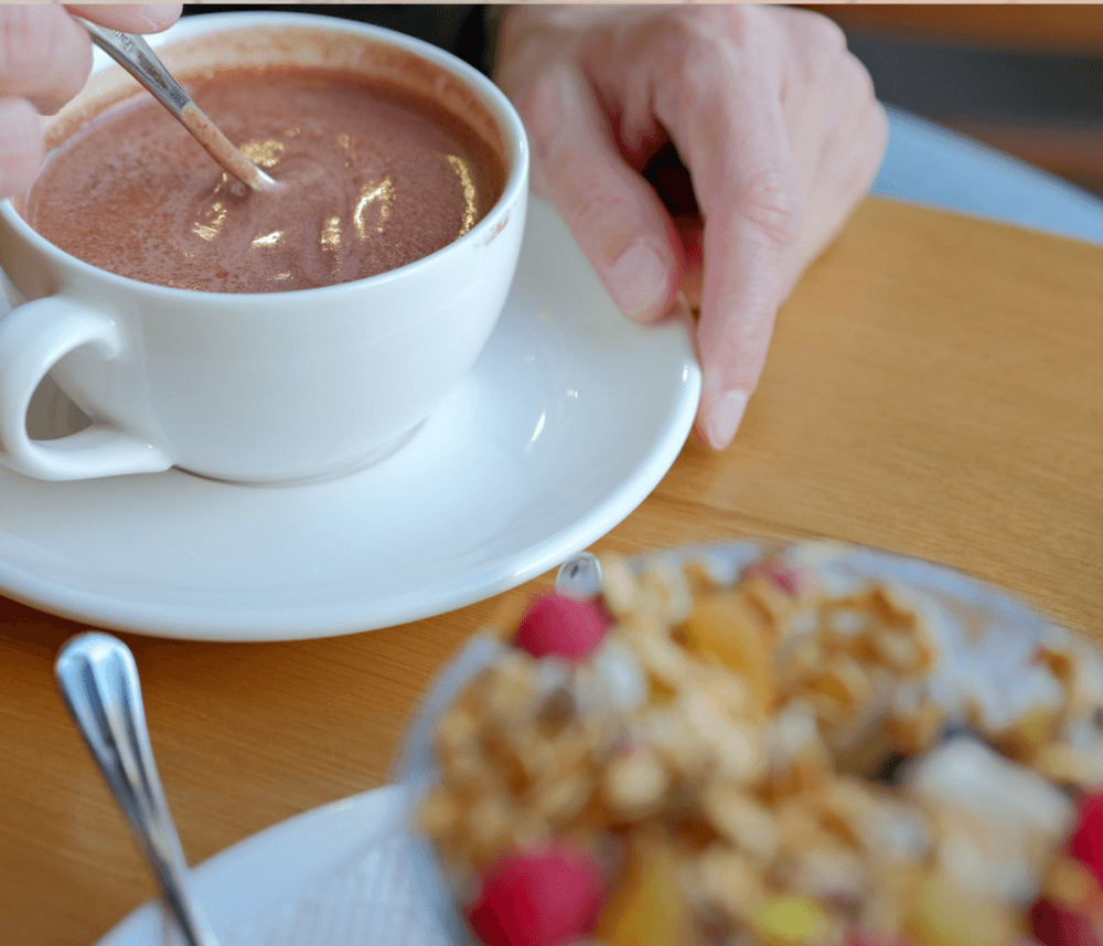 Person stirring a cup of hot chocolate with a spoon, alongside a bowl of granola with fruits. - Home Instead