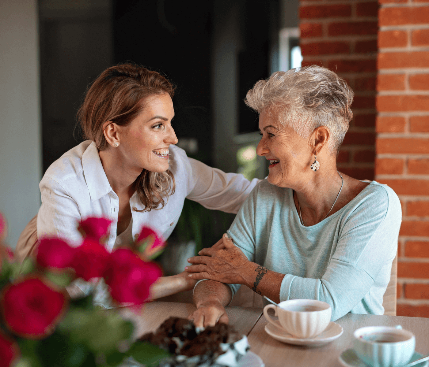 Two women, one younger and one older, smiling and having tea at a table with flowers and a cake. - Home Instead