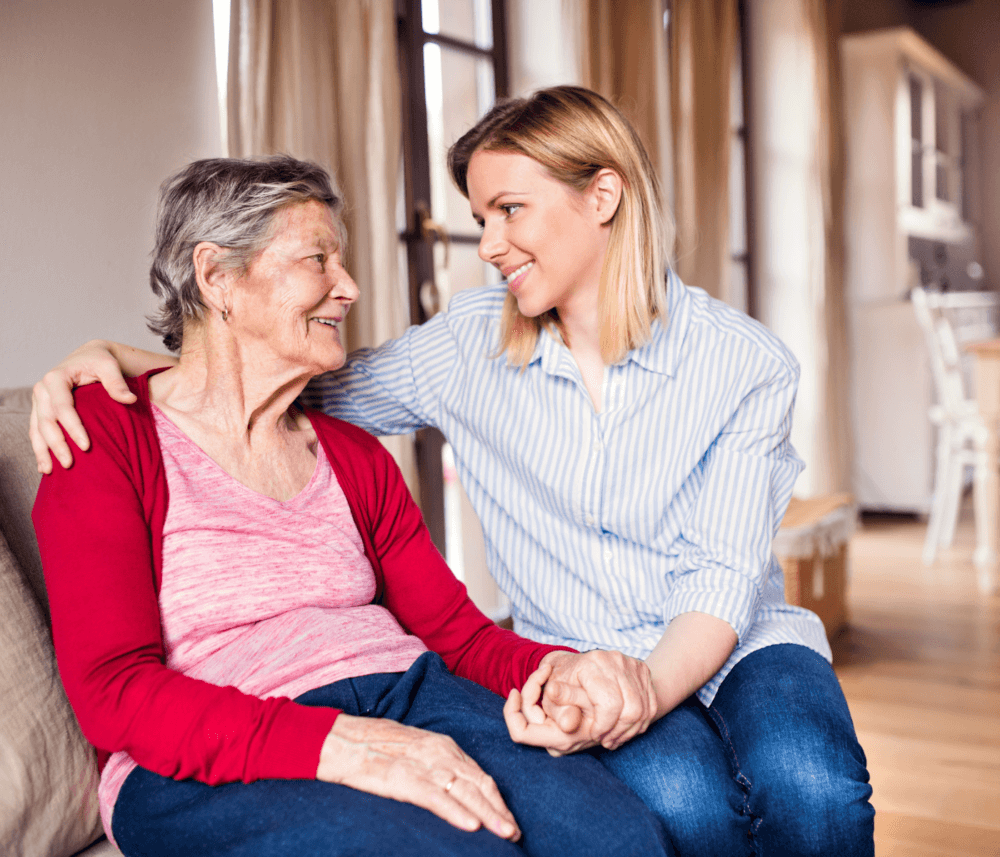 An elderly woman and a younger woman sit together on a couch, smiling and holding hands in a warmly lit room. - Home Instead