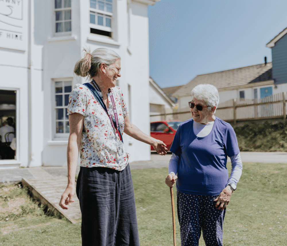 Two women smiling and talking outside a white building on a sunny day, one using a cane and both wearing sunglasses. - Home Instead
