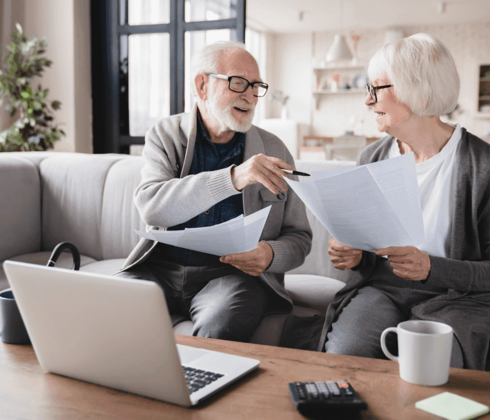 Older couple sitting on a couch, discussing documents, with a laptop and calculator on the coffee table. - Home Instead