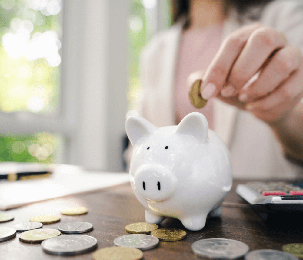 Person placing a coin into a white piggy bank on a table with scattered coins and a calculator nearby. - Home Instead