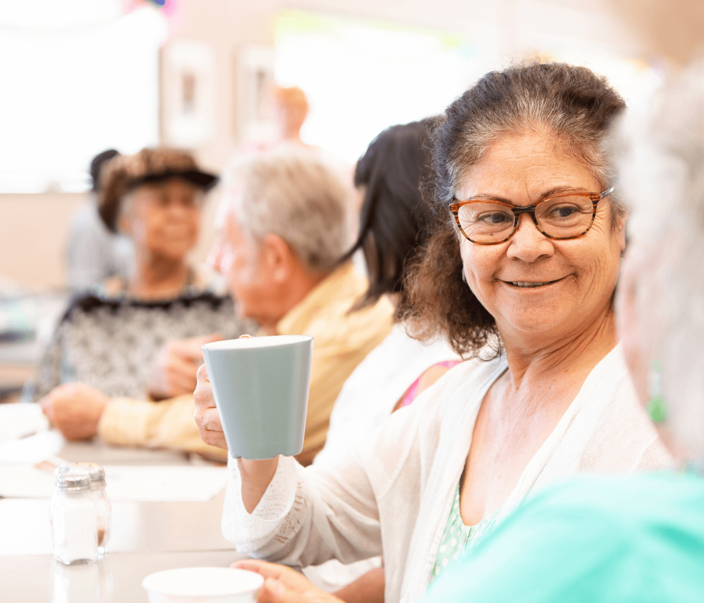 A smiling elderly woman with glasses holds a mug, sitting at a table with other seniors in a social setting. - Home Instead