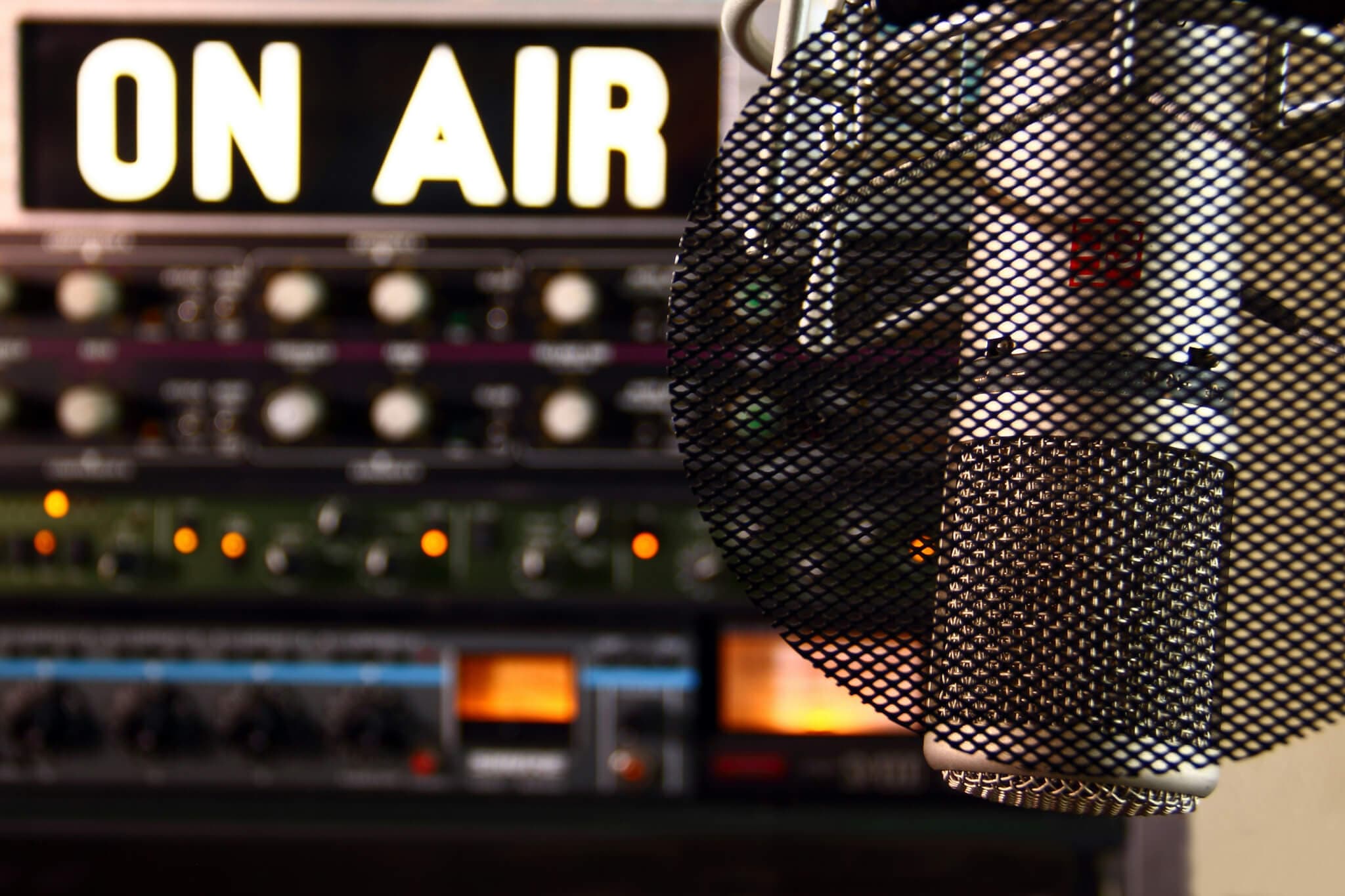Close-up of a studio microphone with a pop filter in front of a blurred "ON AIR" sign and audio equipment. - Home Instead