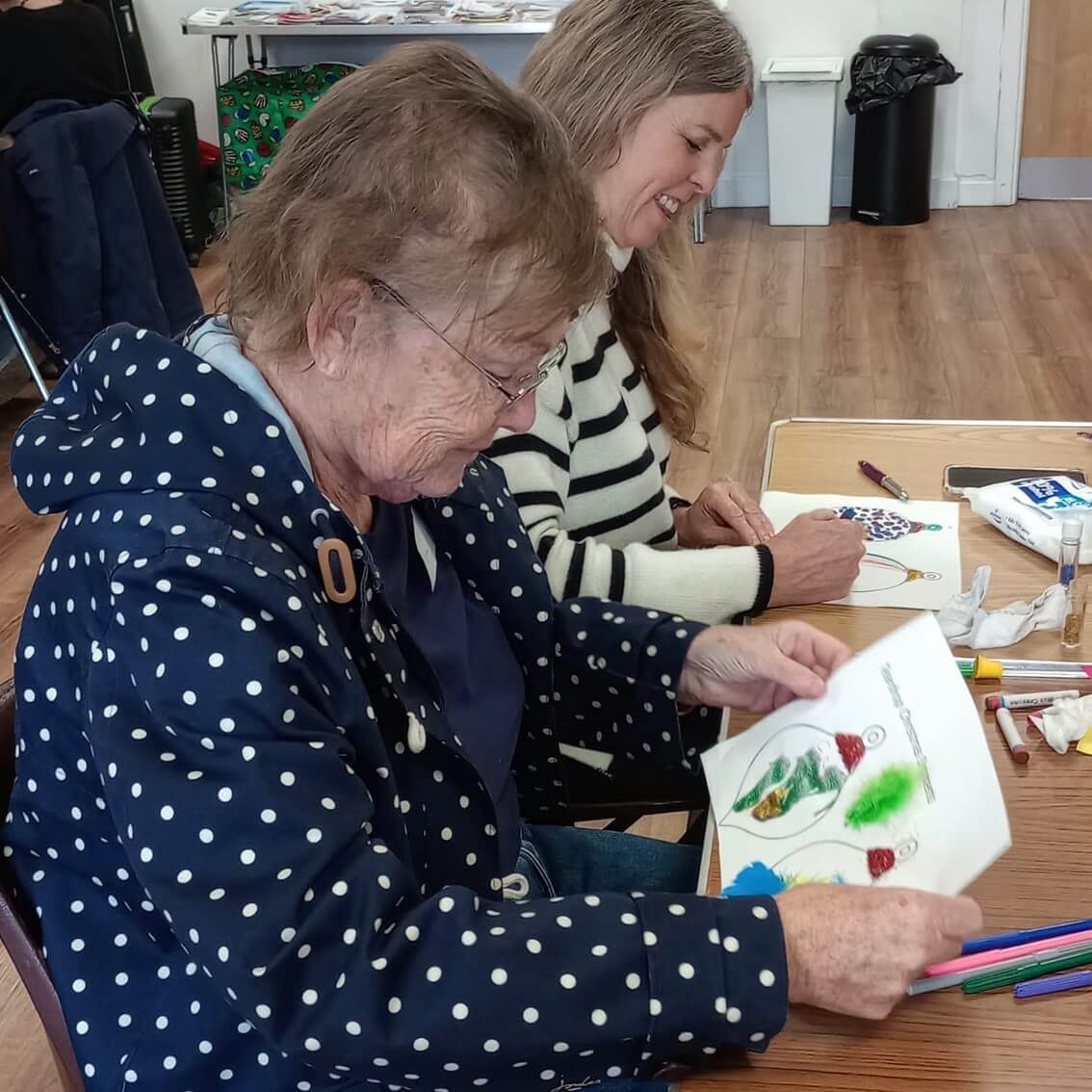Two women sit at a table, smiling and drawing in a bright room, with various art supplies around them. - Home Instead