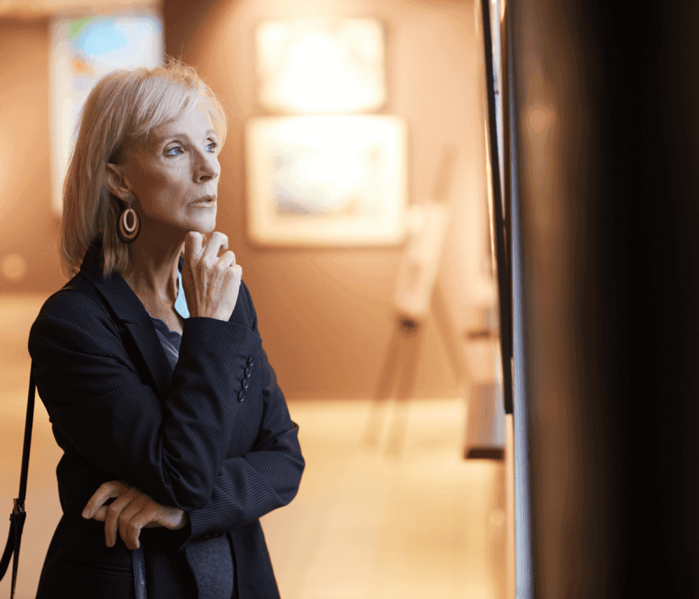 An elderly woman thoughtfully observes artwork in a gallery, with paintings blurred in the background. - Home Instead