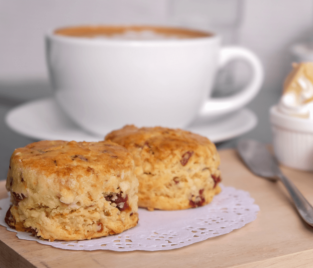 Two scones on a doily with a cup of coffee in the background. A knife and a small pot of cream are on the wooden board. - Home Instead