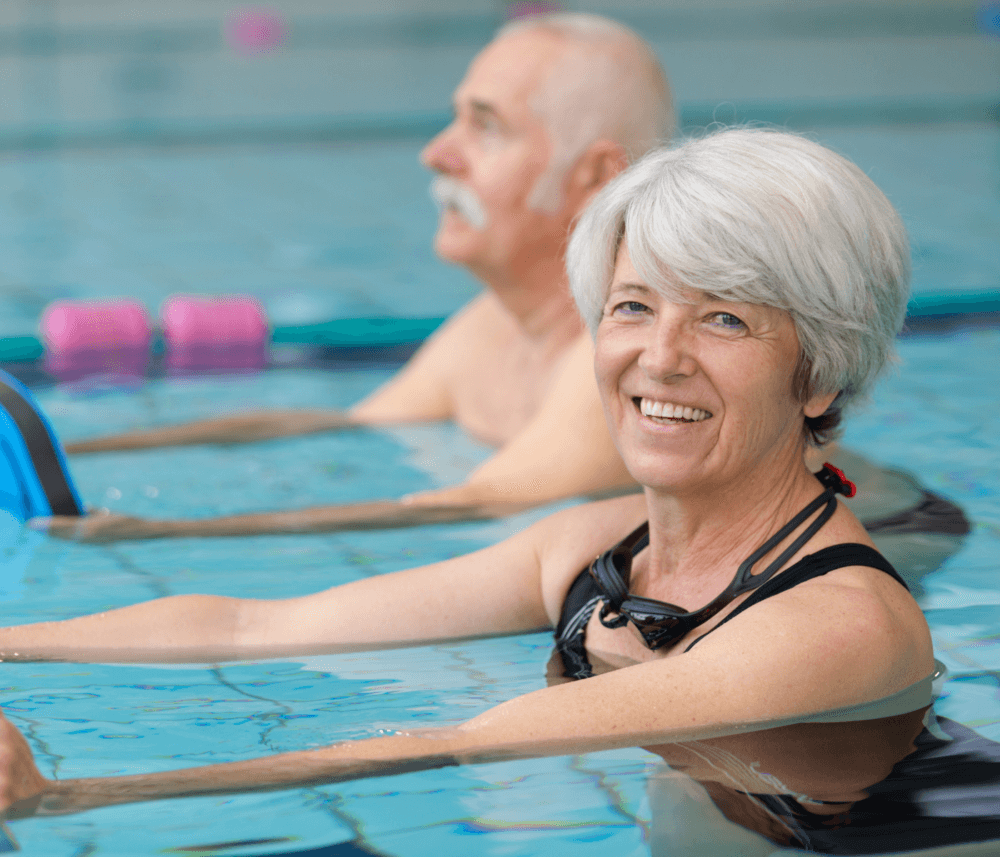 Two smiling older adults in a swimming pool, wearing swimsuits and doing water exercises with foam noodles. - Home Instead