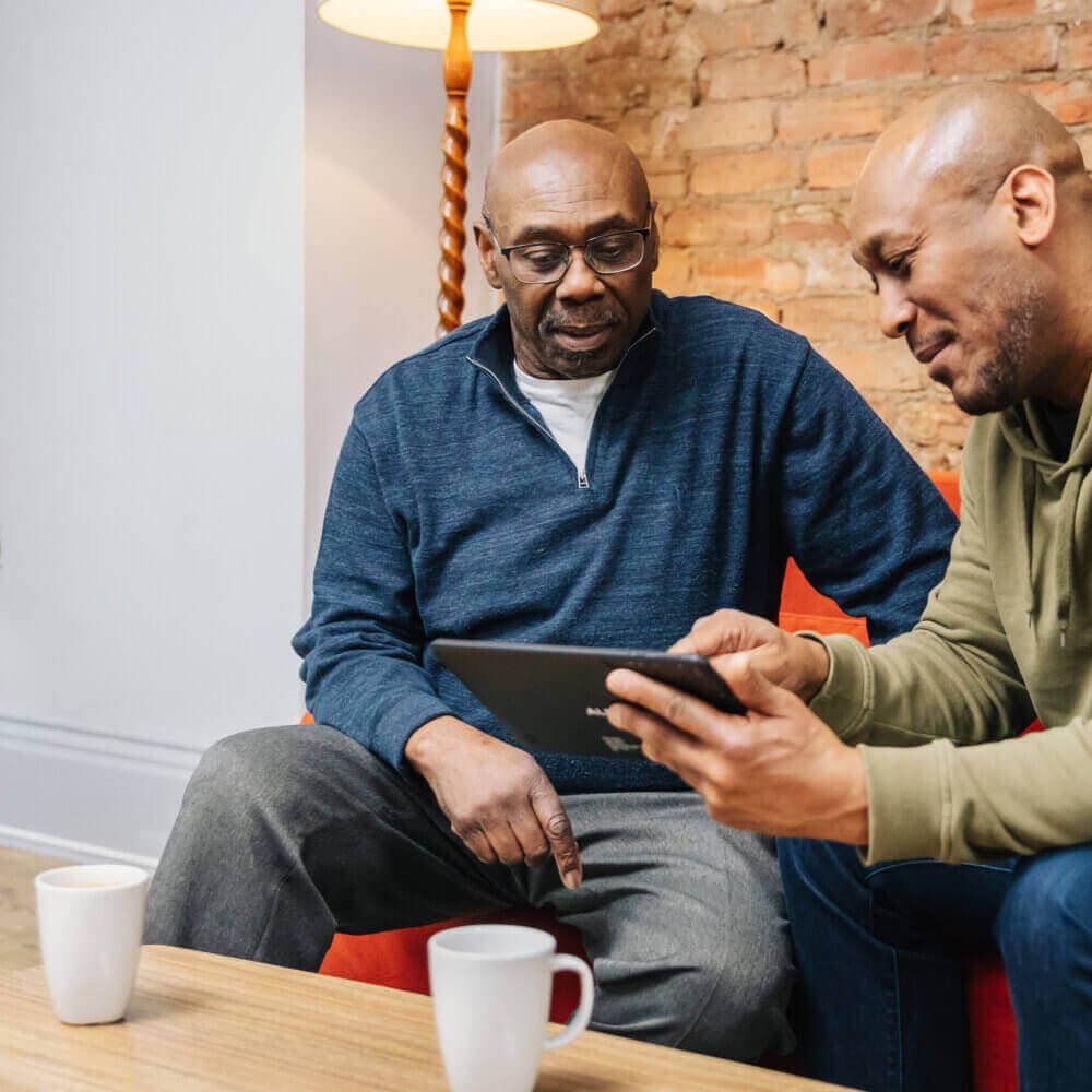Two men sit on a couch with mugs nearby, one showing something on a tablet to the other in a cozy room setting. - Home Instead