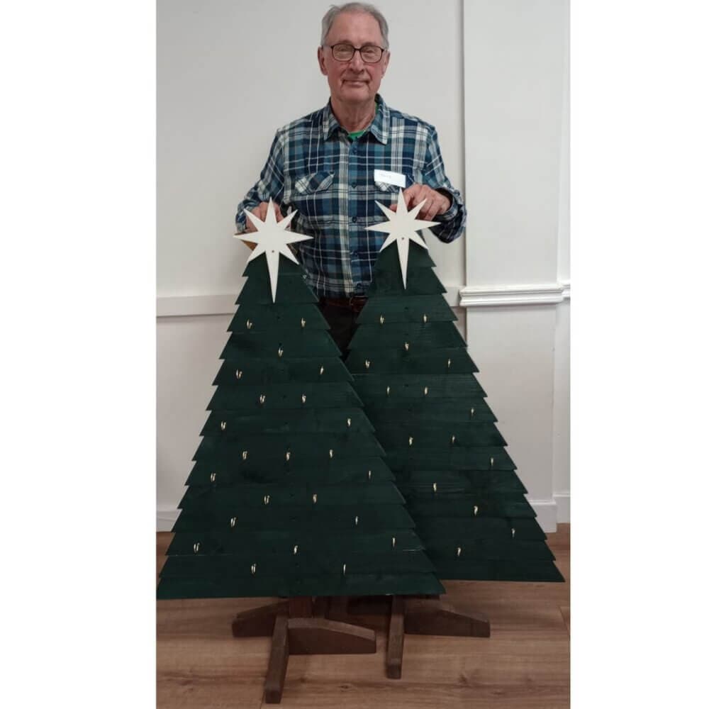 Man in plaid shirt holding two wooden Christmas trees with star toppers in a white-walled room. - Home Instead