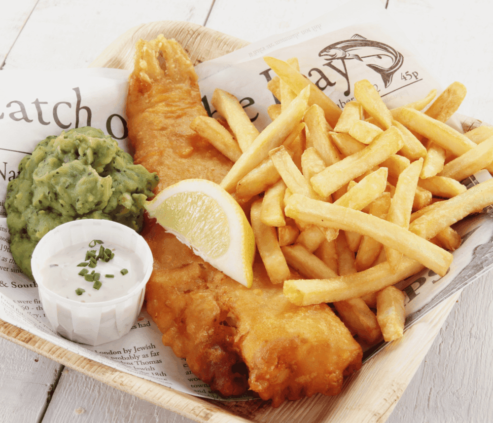 A plate of fish and chips with a lemon wedge, mushy peas, and tartar sauce, served on a newspaper-style wrapper. - Home Instead