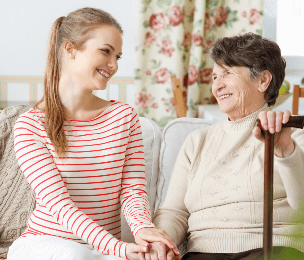 A young woman and an older woman sit on a couch, smiling at each other while holding hands. - Home Instead