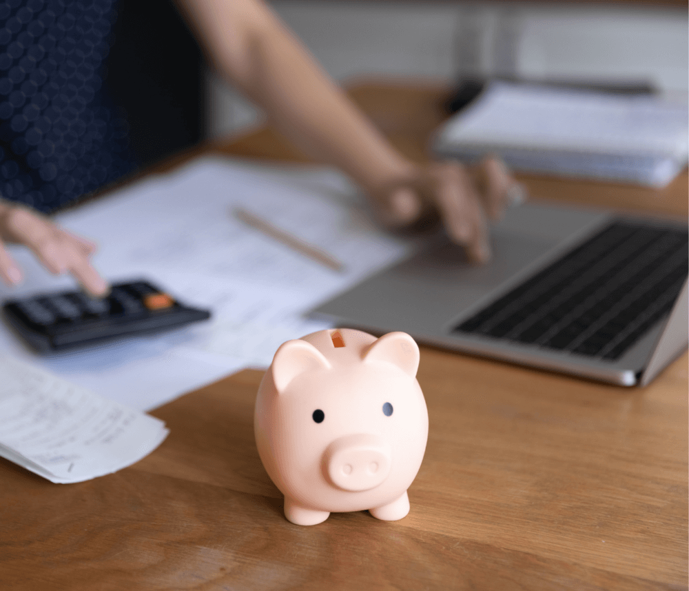 Person working with a calculator and laptop at a desk, with a piggy bank in the foreground. - Home Instead