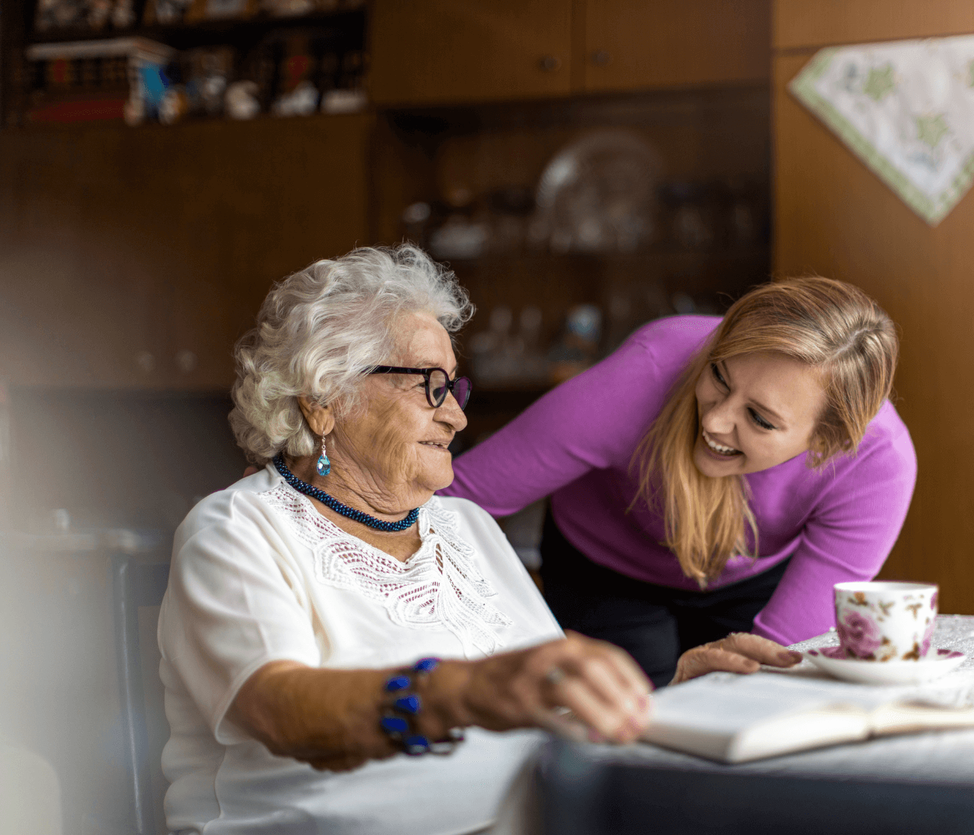 Elderly woman with white hair and glasses sits at table with book, smiling and looking at younger woman in pink sweater. - Home Instead
