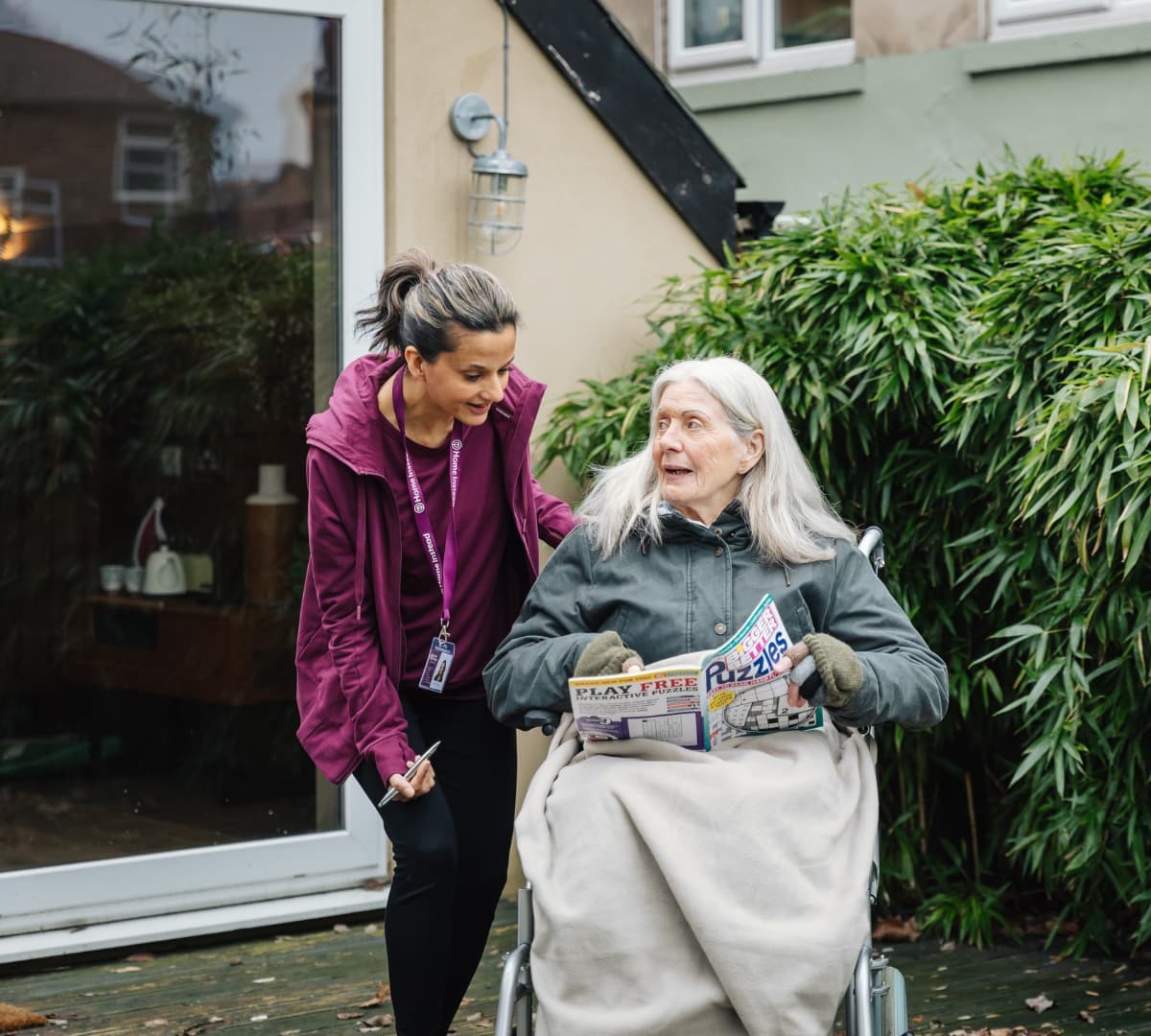 A Care Professional speaks to an elderly woman in a wheelchair holding a magazine outside a house. - Home Instead Southampton