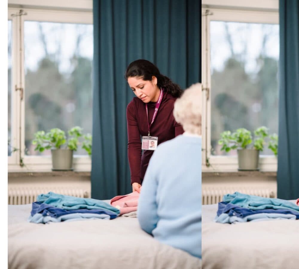 A caregiver helps an elderly person fold clothes in a well-lit room with a window and potted plants. - Home Instead