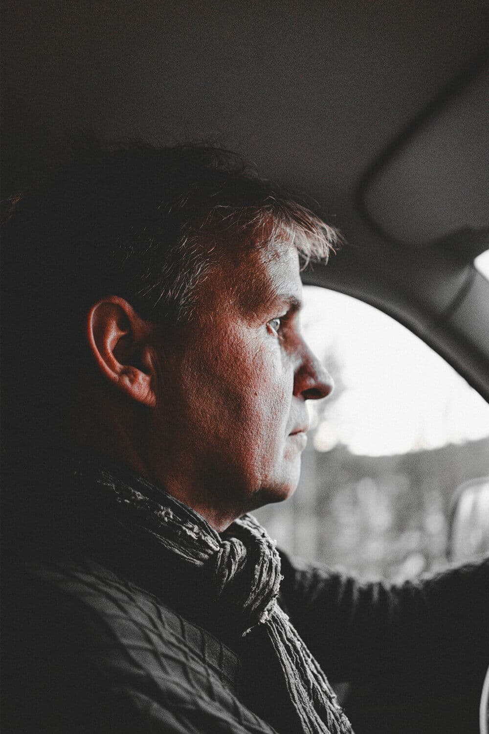 A man with short hair and a scarf drives a vehicle, looking focused and serious. - Home Instead
