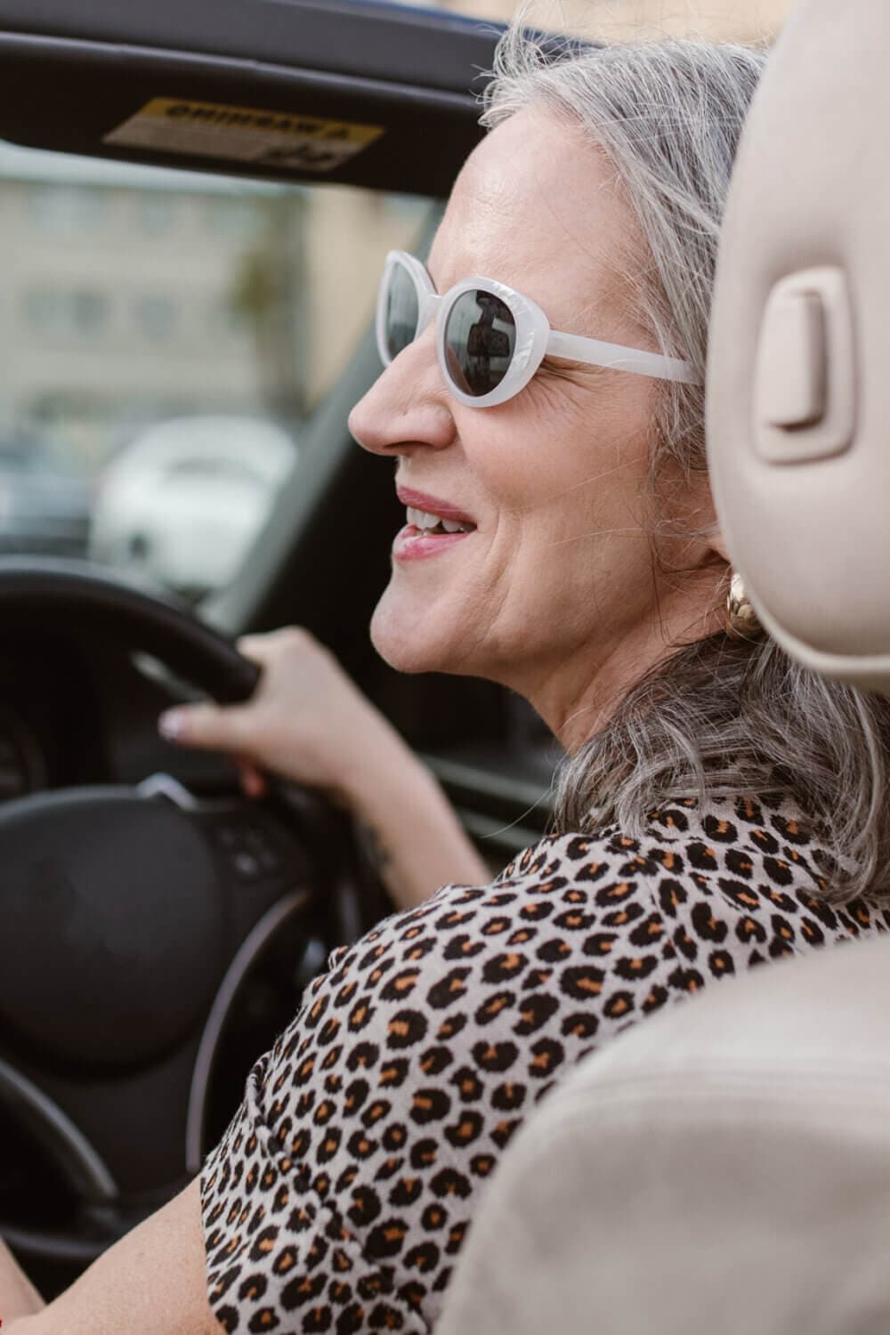 A woman with gray hair and white sunglasses driving a car, smiling, wearing a leopard print shirt. - Home Instead