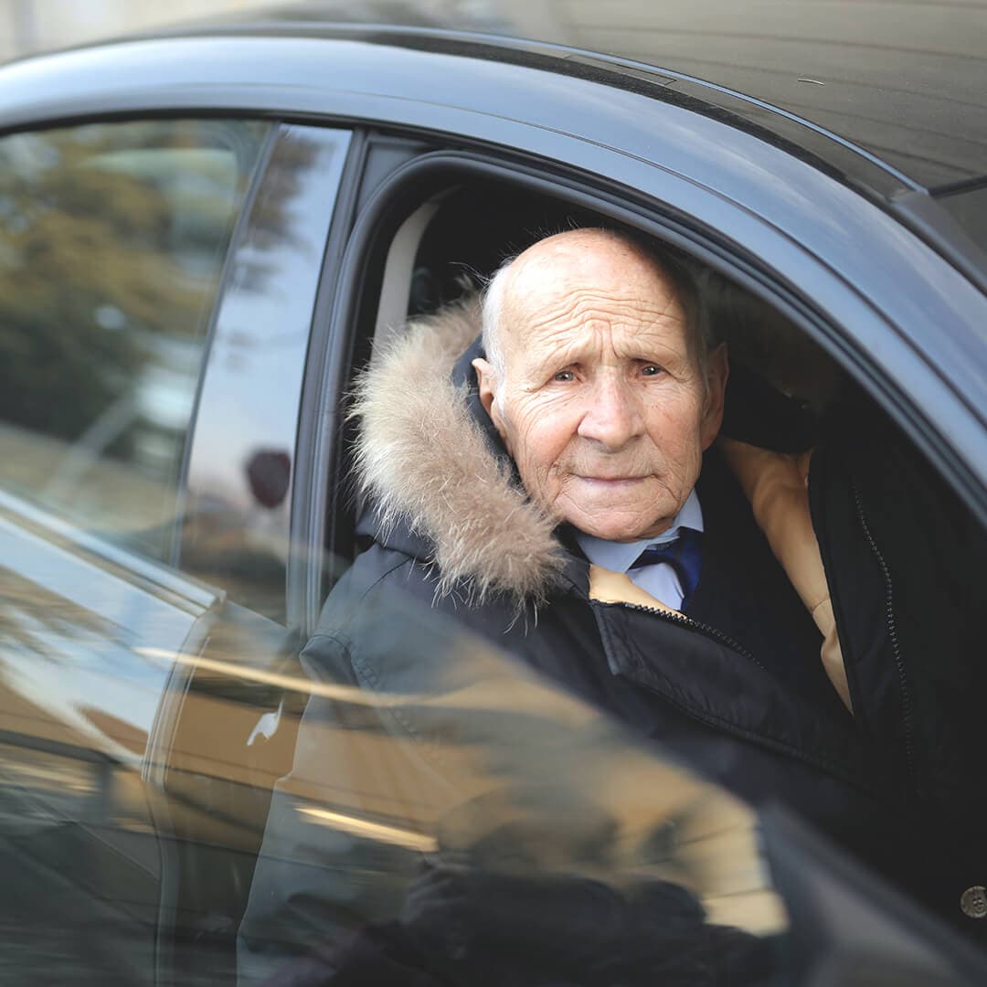 Elderly man in a fur-lined parka sitting in a car, looking out of the open window. - Home Instead