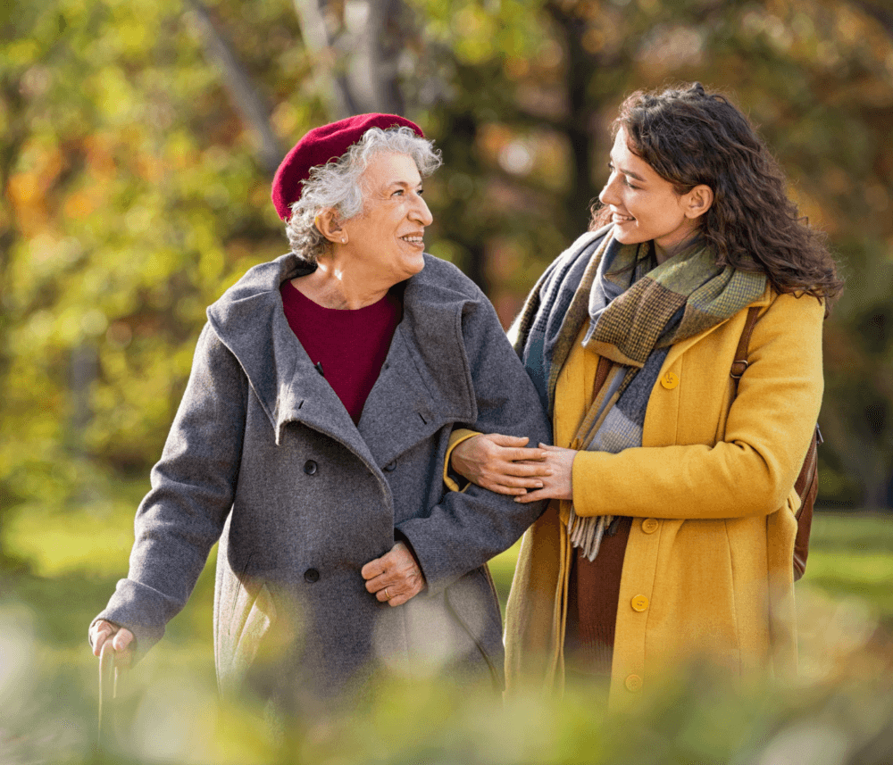 An elderly woman and a younger woman smile at each other while walking arm-in-arm in a park on a sunny day. - Home Instead