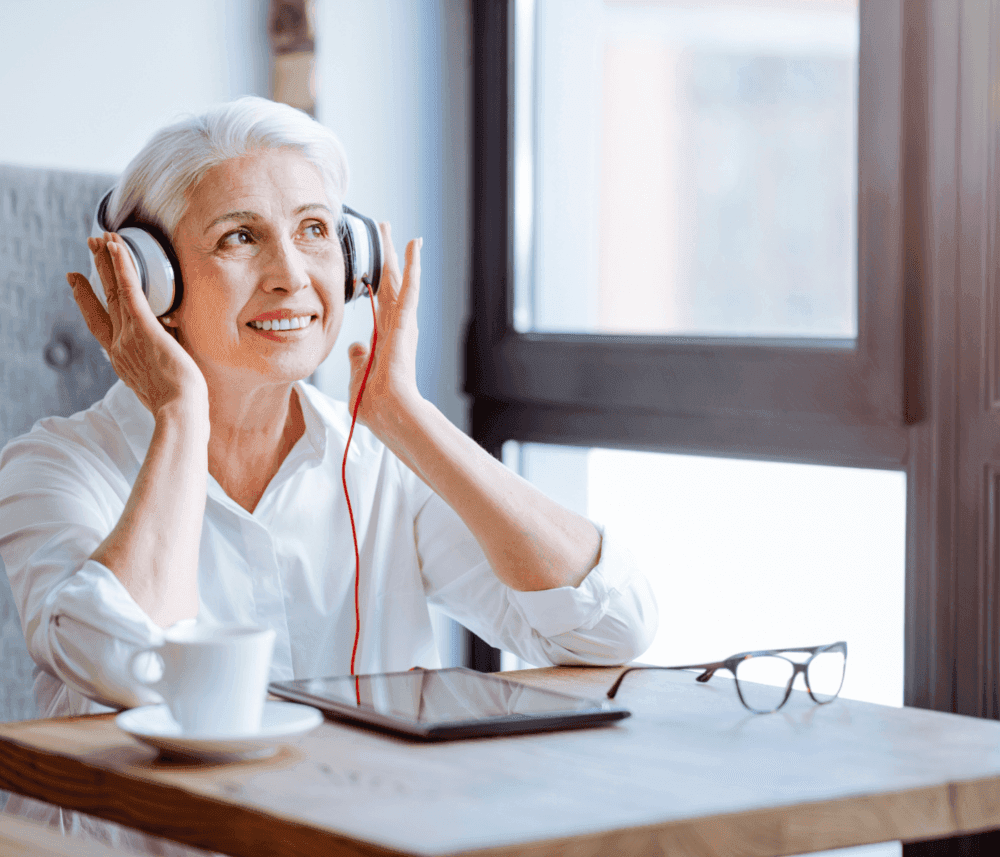 An elderly woman enjoying music through headphones while sitting at a table with a cup of coffee, tablet, and glasses. - Home Instead