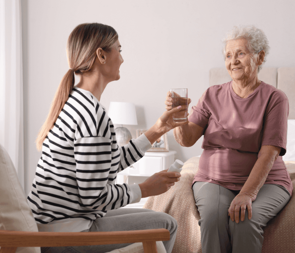 A young woman hands a glass of water to an elderly woman sitting on a bed in a cozy room. - Home Instead