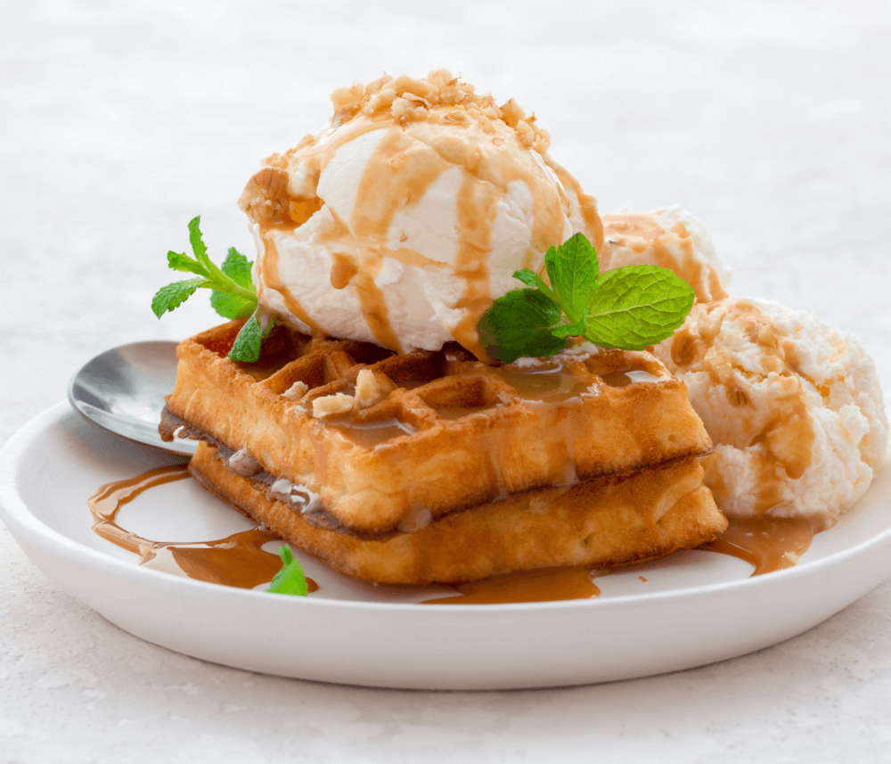 Two waffles topped with ice cream, caramel drizzle, chopped nuts, and mint leaves on a white plate with a spoon. - Home Instead