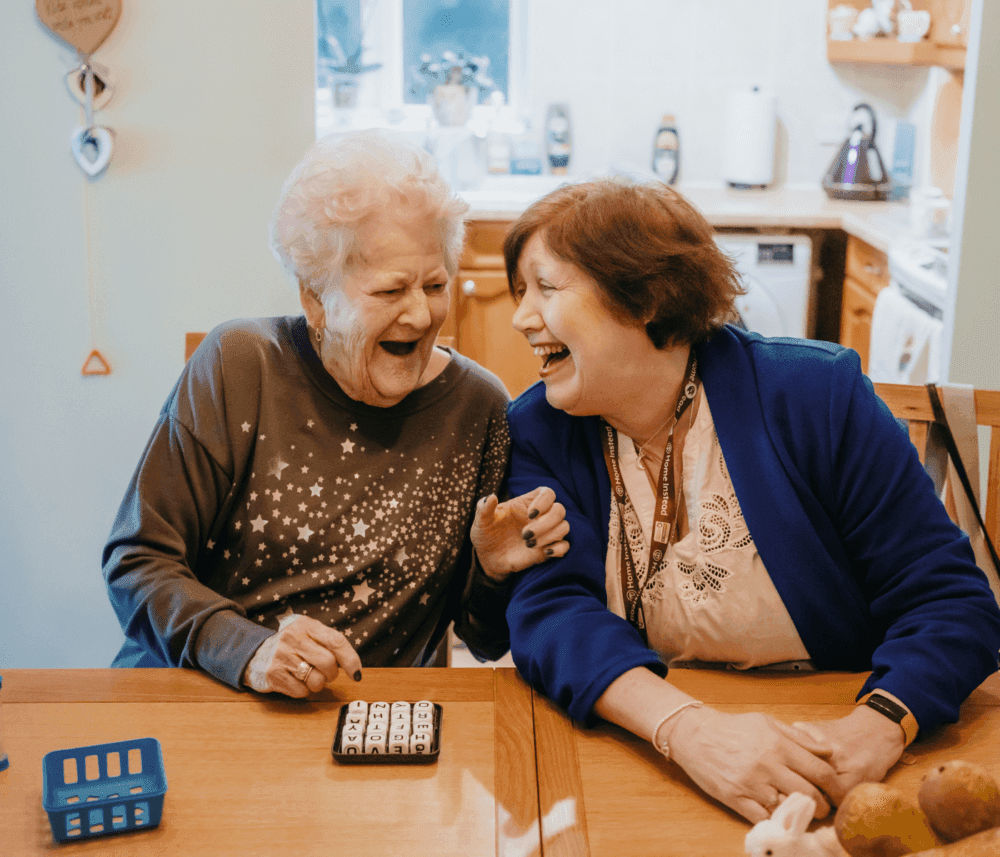 Two women laughing together at a kitchen table, with one pointing at a dice card game. - Home Instead