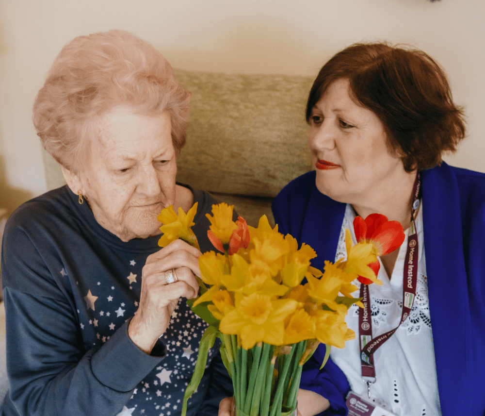 An elderly woman smells yellow flowers while another woman in a blue jacket watches and smiles. - Home Instead