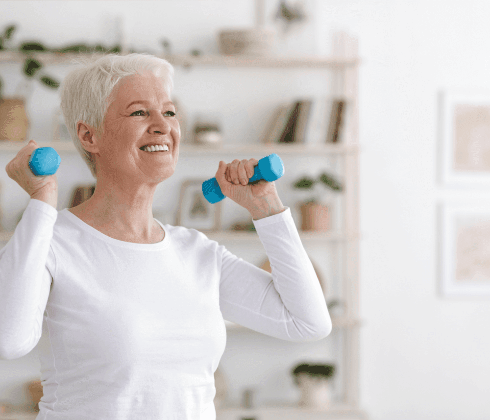 Elderly woman smiling while lifting blue dumbbells for exercise in a bright, well-lit room. - Home Instead