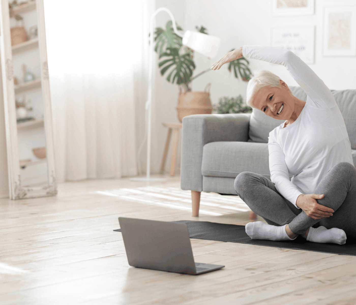 A woman practices yoga on a mat in her living room, smiling and stretching with a laptop in front of her. - Home Instead