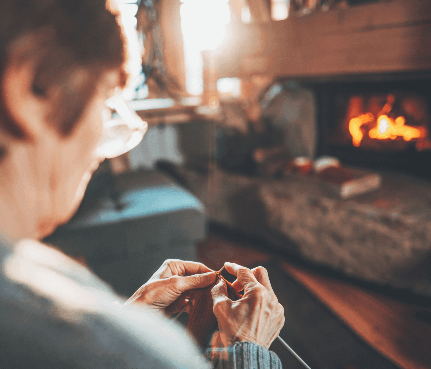 Person knitting by a fireplace in a cozy room, with warm sunlight streaming in and a laid-back atmosphere. - Home Instead