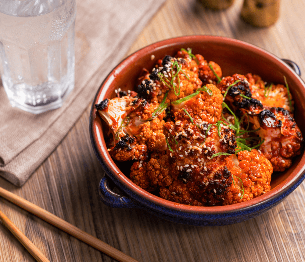 A bowl of roasted cauliflower florets with sesame seeds and green garnish on a wooden table, alongside chopsticks and a glass of water. - Home Instead
