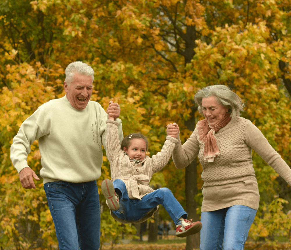 Elderly couple swings a smiling young girl by her arms in a park with colorful autumn leaves in the background. - Home Instead