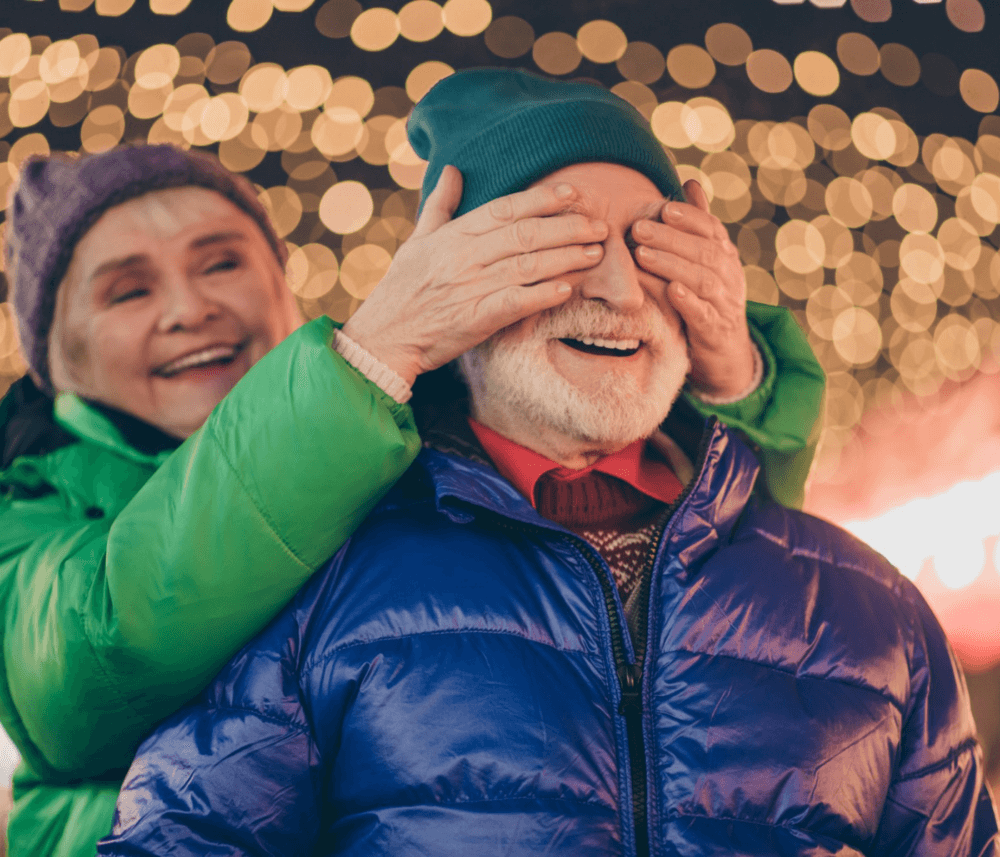 Older woman playfully covers older man's eyes at a festive event with string lights in the background. - Home Instead