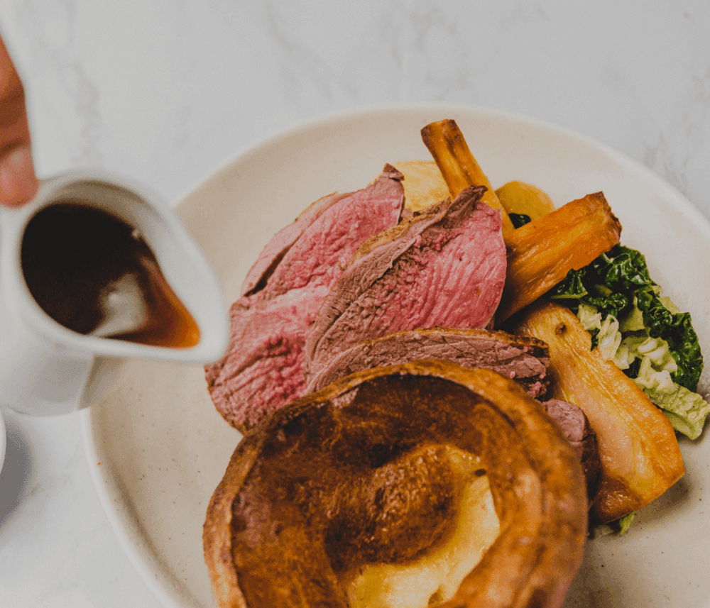 A plate with roast beef, Yorkshire pudding, and vegetables being served with gravy from a gravy boat. - Home Instead