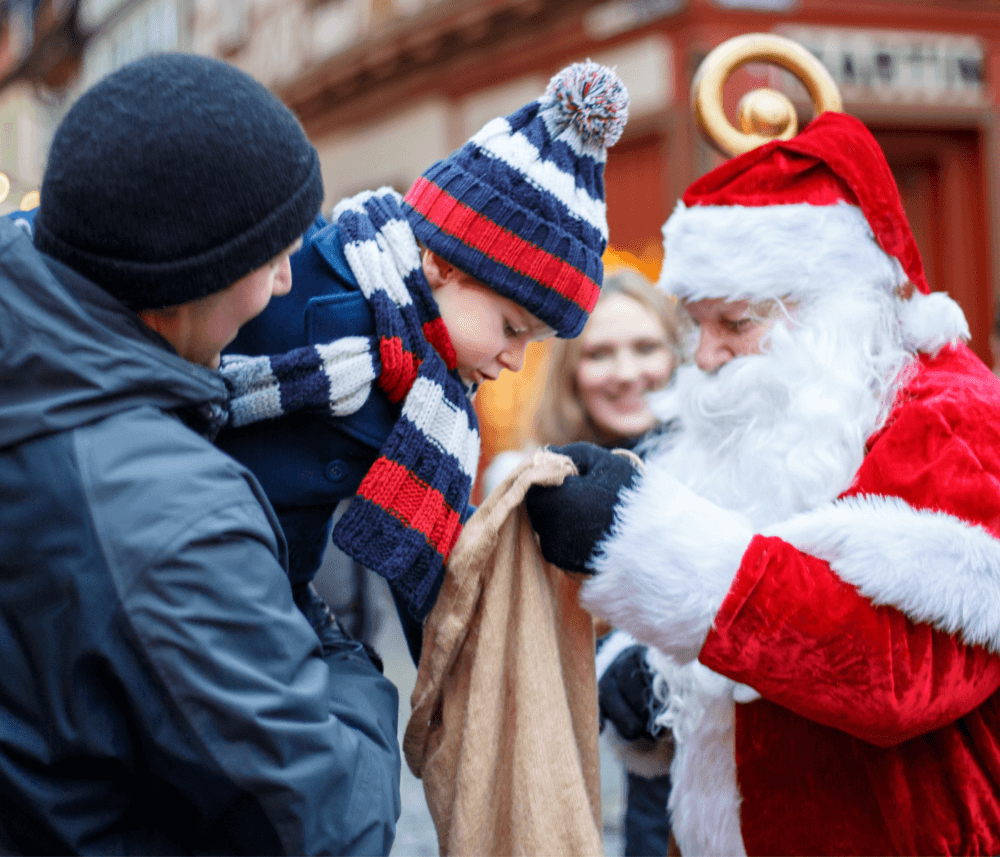 A child held by an adult receives a gift from a person dressed as Santa Claus in an outdoor setting. - Home Instead