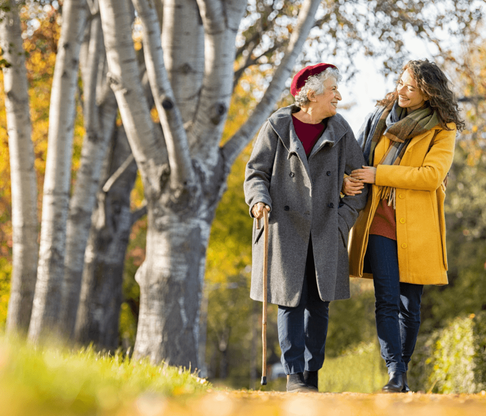 Two women, one older with a cane, walk arm-in-arm on a leafy path among trees in autumn, smiling at one another. - Home Instead