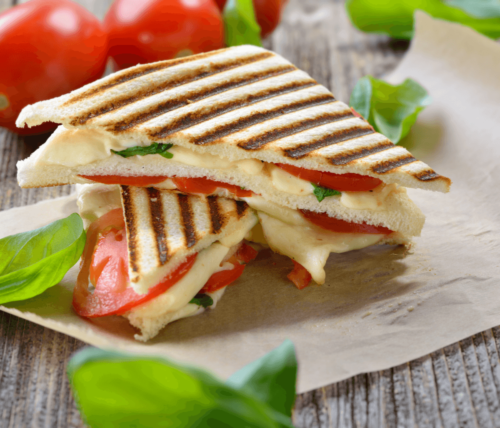 A grilled sandwich with melted cheese, tomatoes, and fresh basil on parchment paper, surrounded by tomatoes and basil leaves. - Home Instead